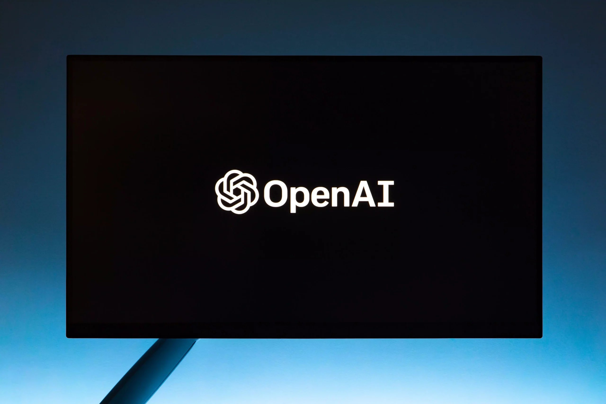 Minimalist display of OpenAI logo on a screen, set against a gradient blue background.