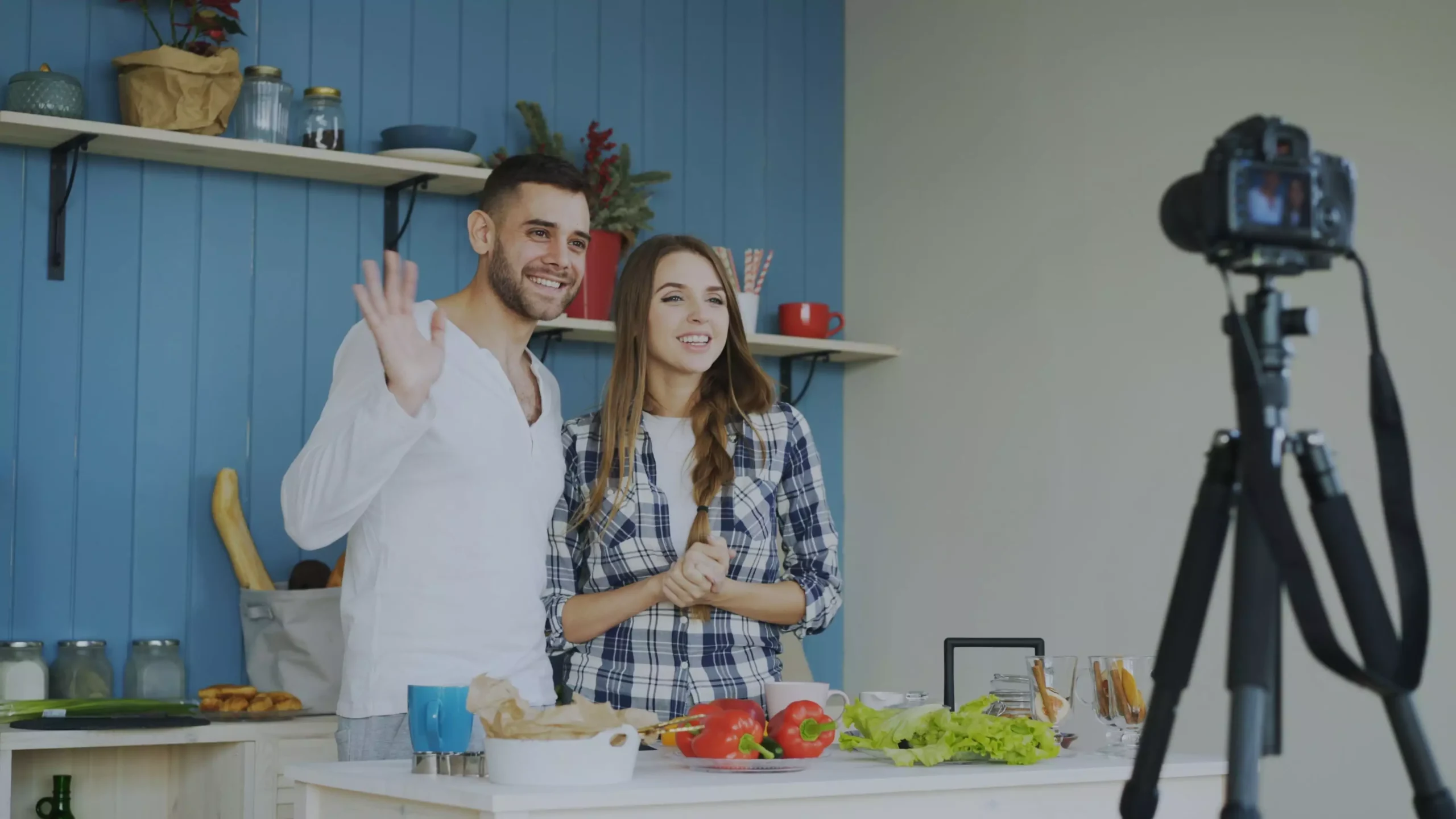 A smiling couple waving and recording a cooking video in their kitchen. Fresh vegetables on the counter.