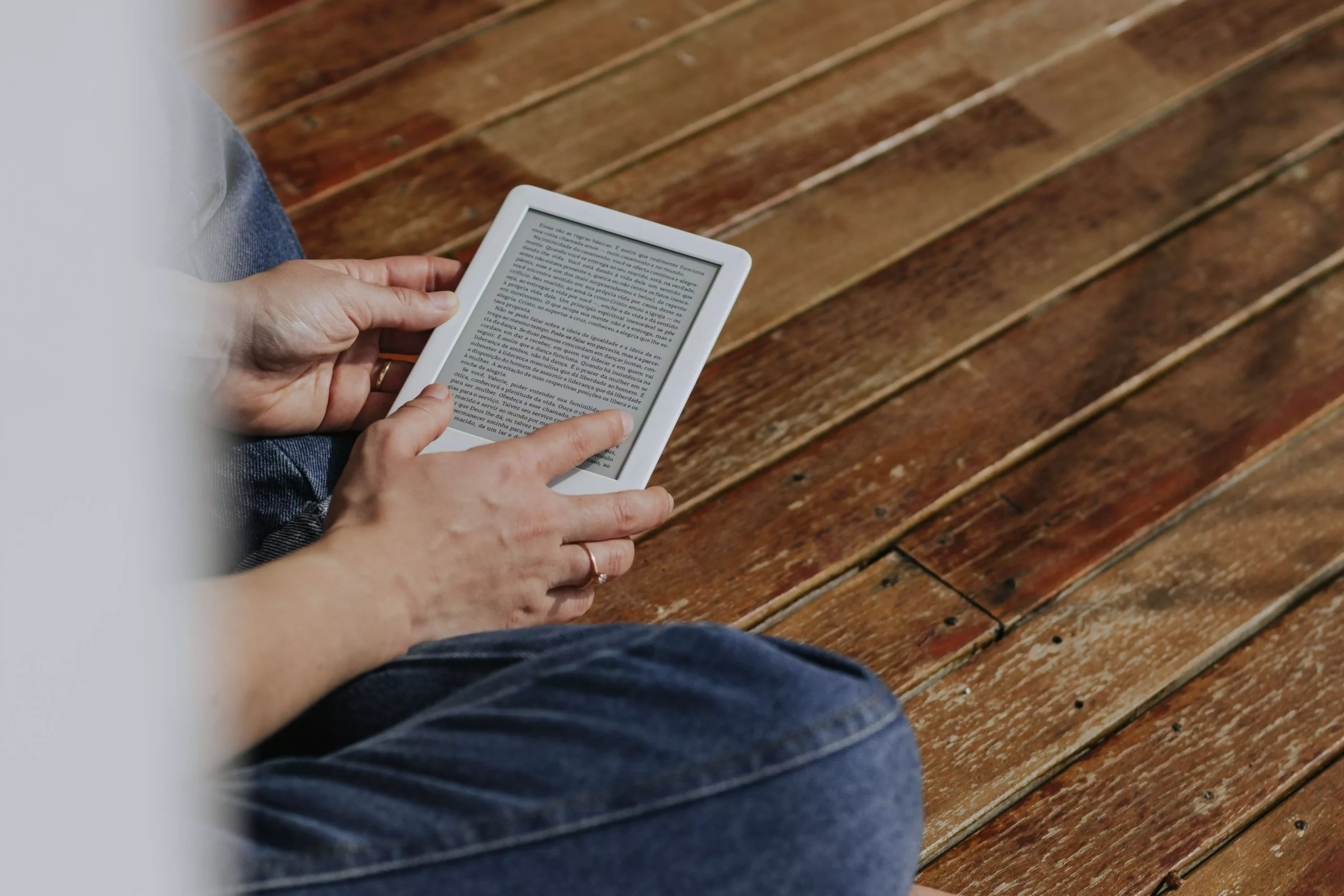 Close-up of hands holding an e-reader on a wooden floor, depicting a casual reading moment.